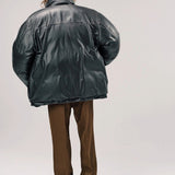 Back view of brown leather puffa jacket on model wearing brown pants and boots, against a white background.