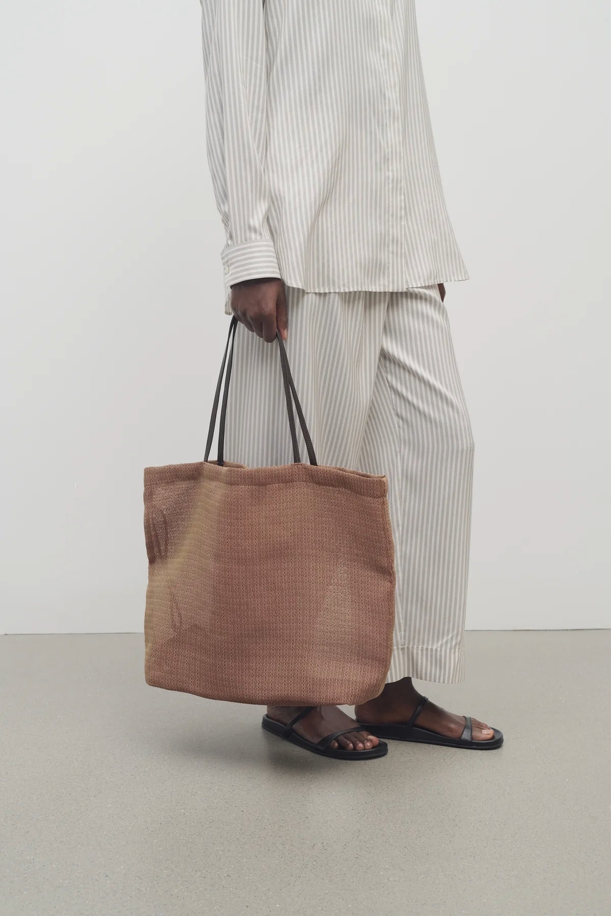 Front view of mesh and leather bag in acero on white background, with model  wearing light silk suit and sandals.