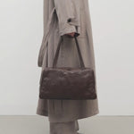 Front view of brown astra bowling bag on model wearing taupe trench coat pants an ballet flats, against a white background.