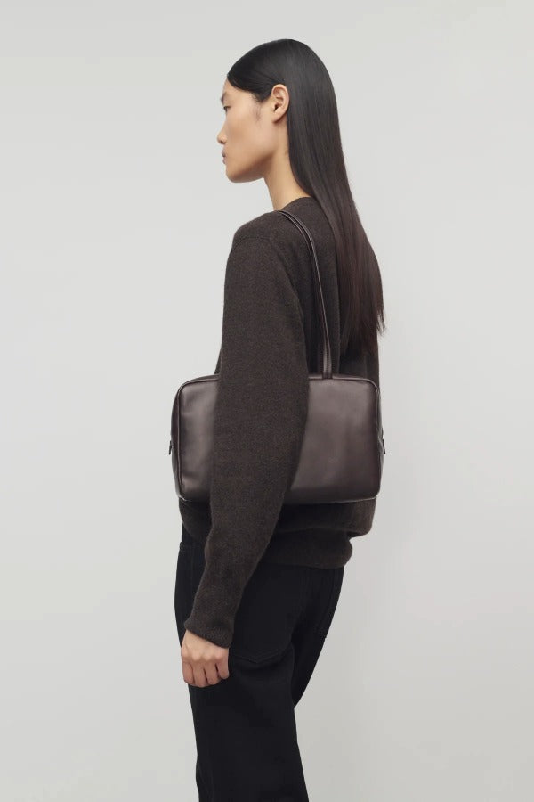 Front view of dark brown astra bowling bag on. model wearing brown sweater and black pants, against a white background.