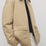Side closeup view of sand aurelius jacket on model wearing black sweater, white shirt and pants, against a white background.