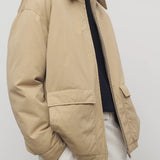 Side closeup view of sand aurelius jacket on model wearing black sweater, white shirt and pants, against a white background.