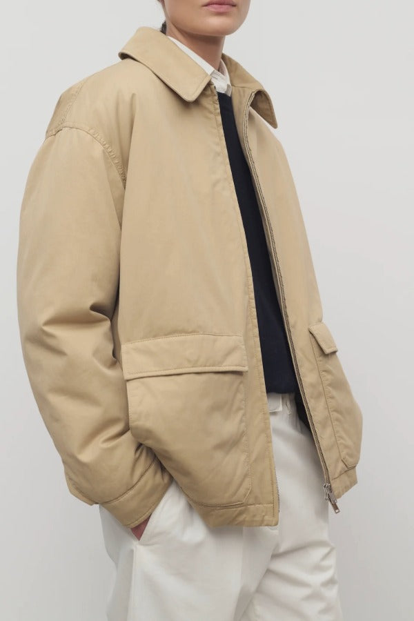 Side closeup view of sand aurelius jacket on model wearing black sweater, white shirt and pants, against a white background.