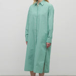 A blonde-haired model is shown in a front-facing view against a white backdrop. She is wearing a long-sleeved, green and white cotton button-down midi dress paired with black sandals.