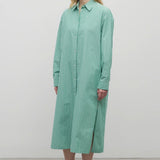 A blonde-haired model is shown in a front-facing view against a white backdrop. She is wearing a long-sleeved, green and white cotton button-down midi dress paired with black sandals.