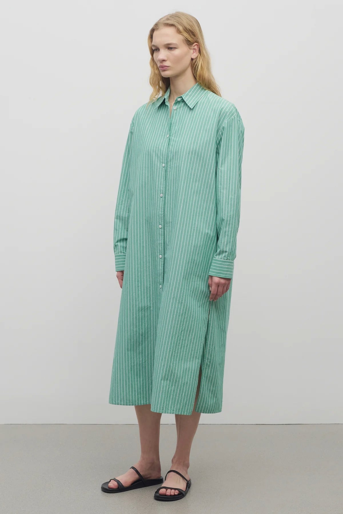 A blonde-haired model is shown in a front-facing view against a white backdrop. She is wearing a long-sleeved, green and white cotton button-down midi dress paired with black sandals.