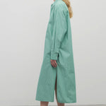 A blonde-haired model is featured in a side-view shot against a plain white backdrop. She is wearing a long-sleeved midi dress made from a green and white cotton fabric, complemented by black sandals.