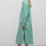 A blonde-haired model is featured in a side-view shot against a plain white backdrop. She is wearing a long-sleeved midi dress made from a green and white cotton fabric, complemented by black sandals.