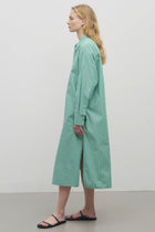 A blonde-haired model is featured in a side-view shot against a plain white backdrop. She is wearing a long-sleeved midi dress made from a green and white cotton fabric, complemented by black sandals.