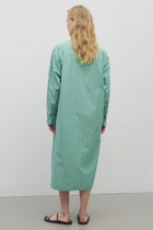 Long blond-haired model wears long-sleeved green with white button-down midi dress and black sandals. Back viewpoint with white background.