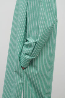 A side closeup of the model wearing a long-sleeved green and white cotton button-down midi dress  is over a white backdrop.