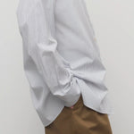 Side view of blue/white cotton sisilia shirt on model wearing camel trousers, against a white background.
