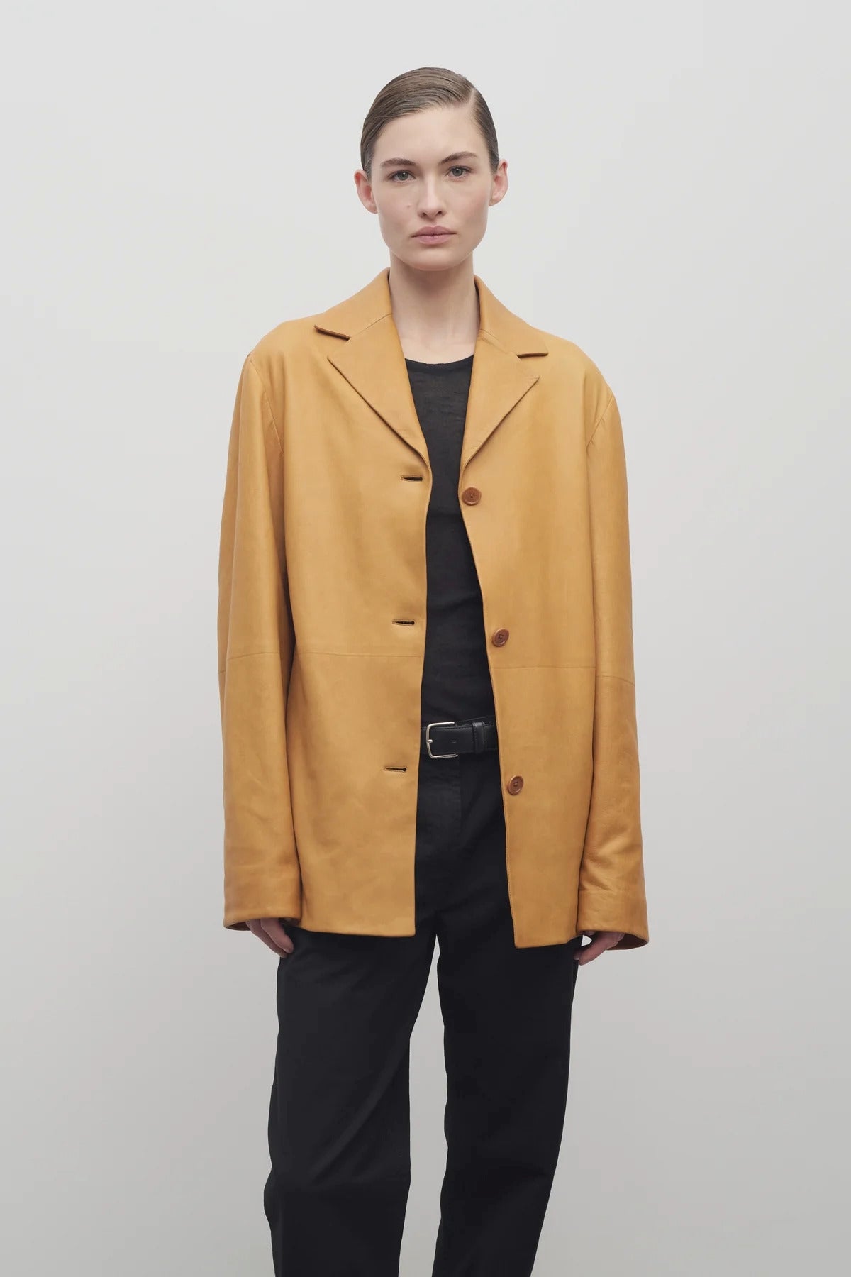 A light brown-haired model is featured wearing the caramel-colored leather button-down jacket, paired with a crew neck black t-shirt, black leather belt, and black trousers. Front viewpoint over white backdrop.