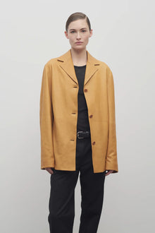 A light brown-haired model is featured wearing the caramel-colored leather button-down jacket, paired with a crew neck black t-shirt, black leather belt, and black trousers. Front viewpoint over white backdrop.