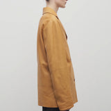 A light brown-haired model is shown in a side view against a white backdrop. She is wearing a caramel-colored leather button-down jacket paired with black trousers.