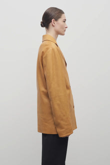 A light brown-haired model is shown in a side view against a white backdrop. She is wearing a caramel-colored leather button-down jacket paired with black trousers.