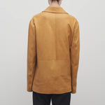 A light brown-haired model is wearing a caramel-colored leather button-down jacket paired with black trousers. The photo is taken from a back viewpoint against a white backdrop.