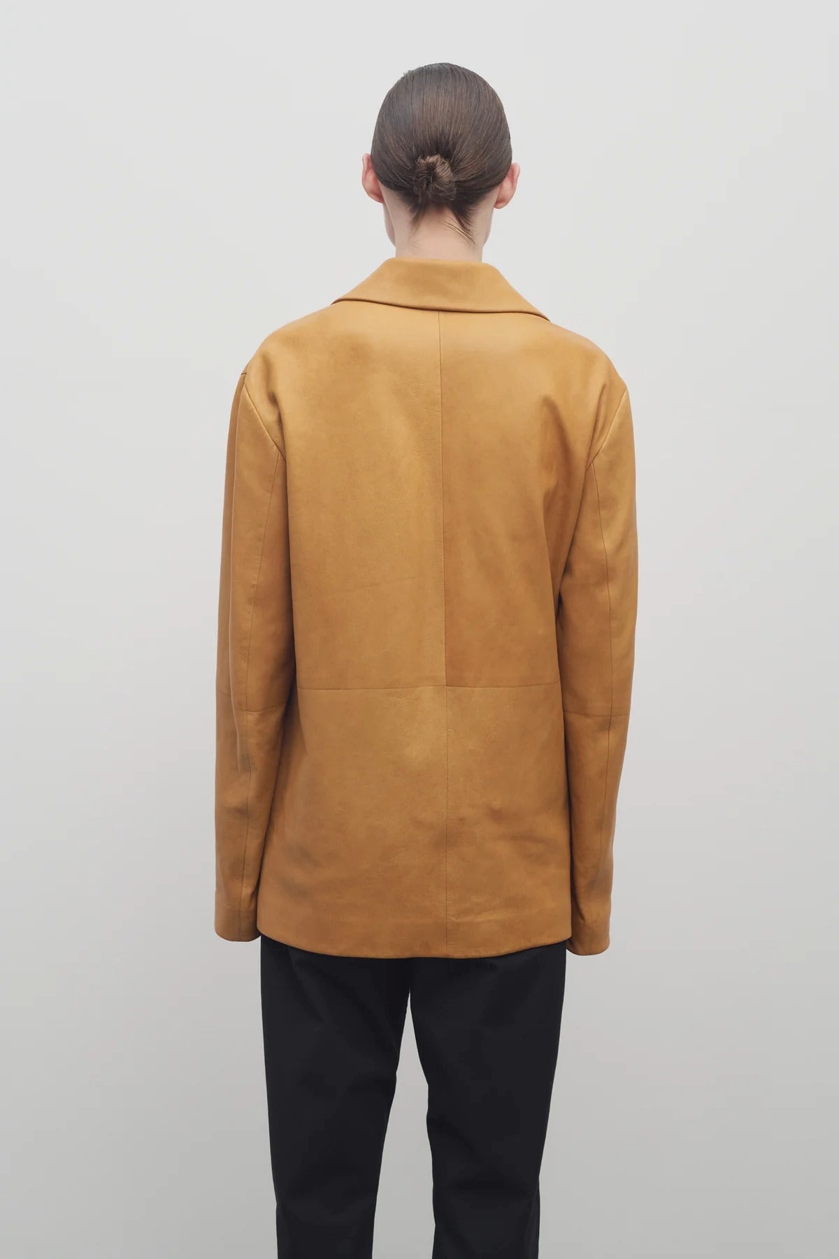 A light brown-haired model is wearing a caramel-colored leather button-down jacket paired with black trousers. The photo is taken from a back viewpoint against a white backdrop.