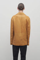 A light brown-haired model is wearing a caramel-colored leather button-down jacket paired with black trousers. The photo is taken from a back viewpoint against a white backdrop.