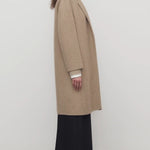 Side view of natural fedra coat on model wearing black skirt and mules, against a white background.