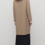 Back view of natural fedra coat on model wearing black skirt and mules, against a white backround.
