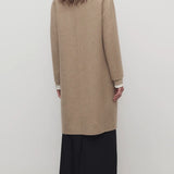 Back view of natural fedra coat on model wearing black skirt and mules, against a white backround.