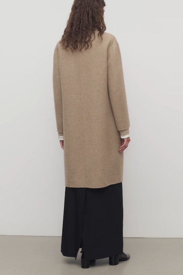 Back view of natural fedra coat on model wearing black skirt and mules, against a white backround.