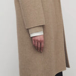 Side closeup view of natural fedra coat on model wearing black skirt, against a white background.