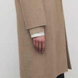 Side closeup view of natural fedra coat on model wearing black skirt, against a white background.