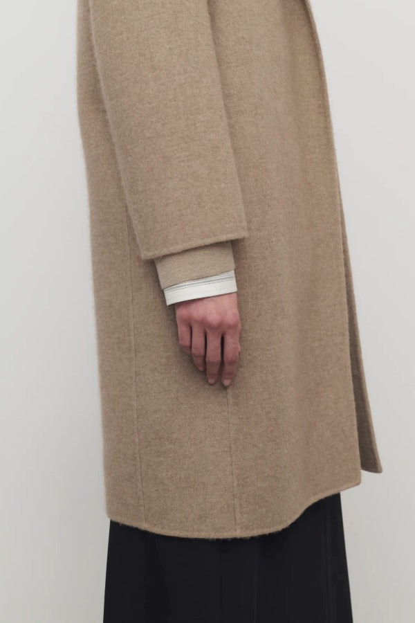 Side closeup view of natural fedra coat on model wearing black skirt, against a white background.