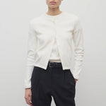 A model with dark brown hair is shown from the front, set against a white backdrop. The model is wearing a long-sleeved cotton button-down sweater over a white crew neck t-shirt. The outfit is completed with black trousers and a classic black belt.