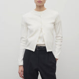 A model with dark brown hair is shown from the front, set against a white backdrop. The model is wearing a long-sleeved cotton button-down sweater over a white crew neck t-shirt. The outfit is completed with black trousers and a classic black belt.