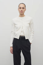 A model with dark brown hair is shown from the front, set against a white backdrop. The model is wearing a long-sleeved cotton button-down sweater over a white crew neck t-shirt. The outfit is completed with black trousers and a classic black belt.