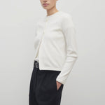 A dark brown-haired model is showcasing a long-sleeved white cotton button-down sweater, layered over a white crew-neck t-shirt. The look is completed with a classic black belt and black trousers.