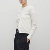 A dark brown-haired model is showcasing a long-sleeved white cotton button-down sweater, layered over a white crew-neck t-shirt. The look is completed with a classic black belt and black trousers.