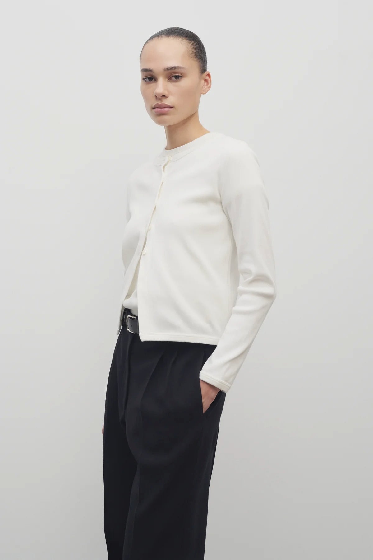 A dark brown-haired model is showcasing a long-sleeved white cotton button-down sweater, layered over a white crew-neck t-shirt. The look is completed with a classic black belt and black trousers.