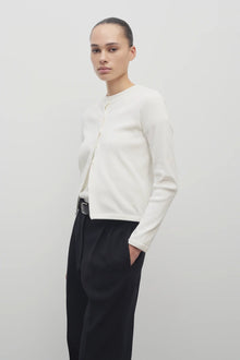 A dark brown-haired model is showcasing a long-sleeved white cotton button-down sweater, layered over a white crew-neck t-shirt. The look is completed with a classic black belt and black trousers.