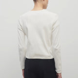 A dark brown-haired model is featured wearing a long-sleeved white cotton button-down sweater paired with black trousers. The viewpoint is from the back, and the background is a plain white backdrop.