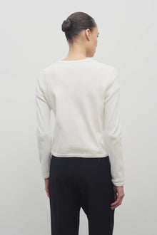 A dark brown-haired model is featured wearing a long-sleeved white cotton button-down sweater paired with black trousers. The viewpoint is from the back, and the background is a plain white backdrop.