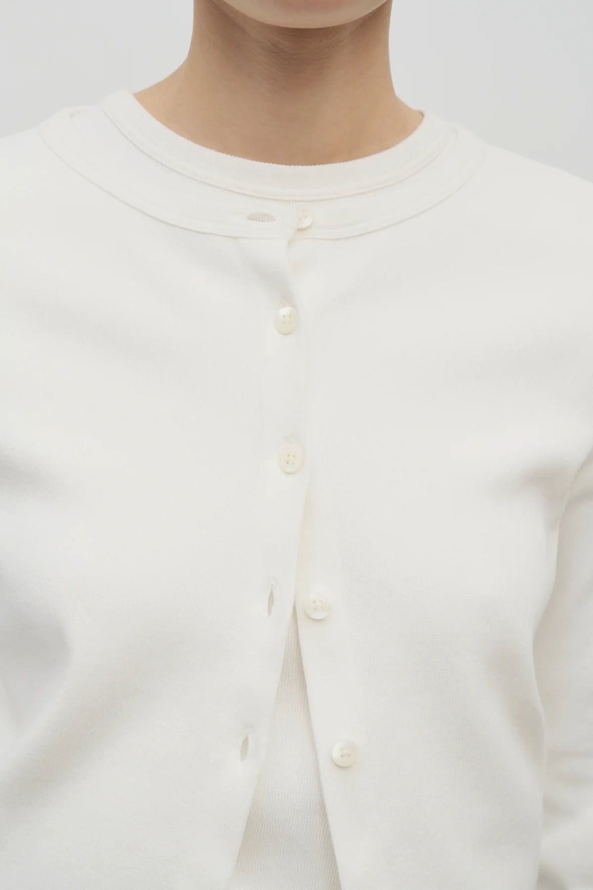 A model wears a long-sleeved white cotton button-down cardigan over a crew neck white t-shirt. The viewpoint is a closeup of the model from the front.
