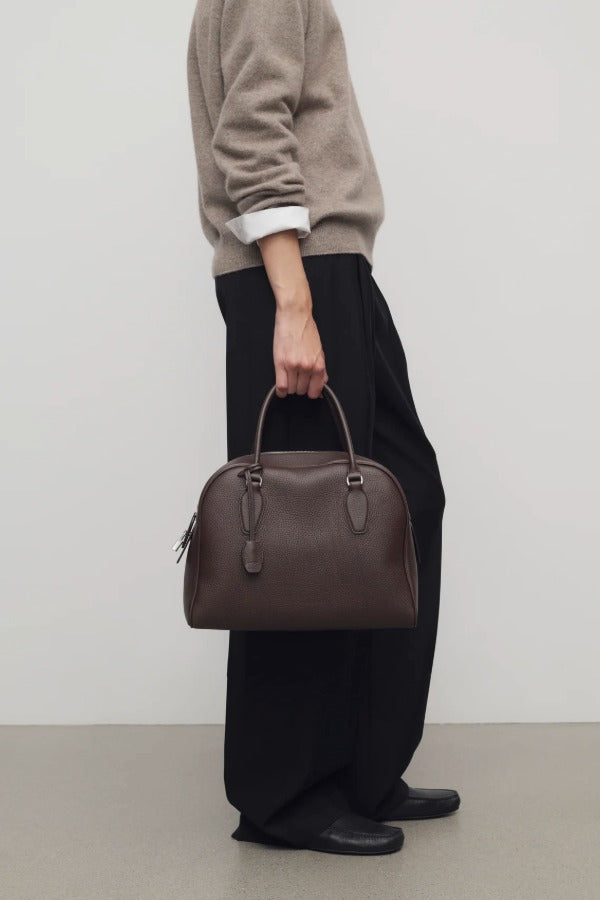 Front view of deep brown india 12 bag on model wearing black pants, taupe sweater and loafers, against a white background.