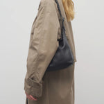 Back side of black isa shoulder bag on model wearing khaki trench coat, against white background.