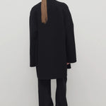 Back view of black kala coat on model wearing black turtleneck and trousers, against a white background.