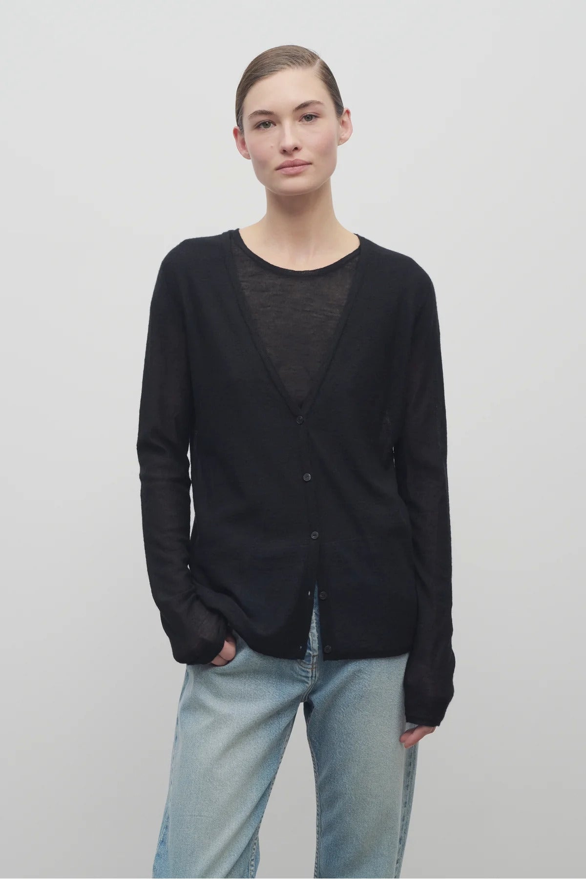 A model with light brown hair is shown wearing the black long sleeve knit cardigan with button down closure over a crew neck t-shirt and baggy jeans. Viewpoint is frontside with a white backdrop.