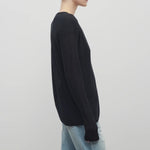 A model with light brown hair wears a long sleeve black knit cardigan with baggy jeans shown at a side angle against a white backdrop.