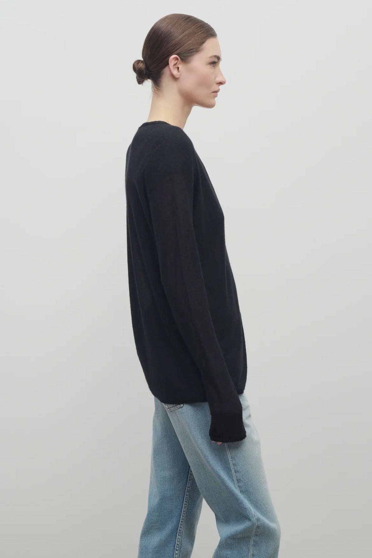 A model with light brown hair wears a long sleeve black knit cardigan with baggy jeans shown at a side angle against a white backdrop.