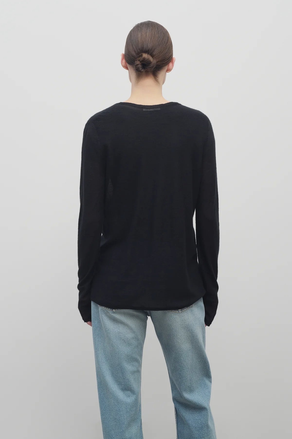 A model with light brown hair wears the long sleeve black knit cardigan with baggy jeans shown backside with a white backdrop.