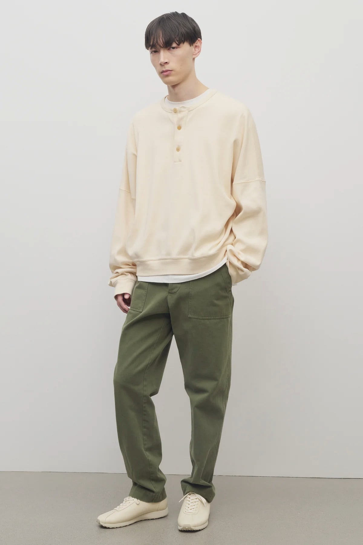 Front slanted view of khaki green pants on model wearing white button down sweatshirt and white shoes, against white background.