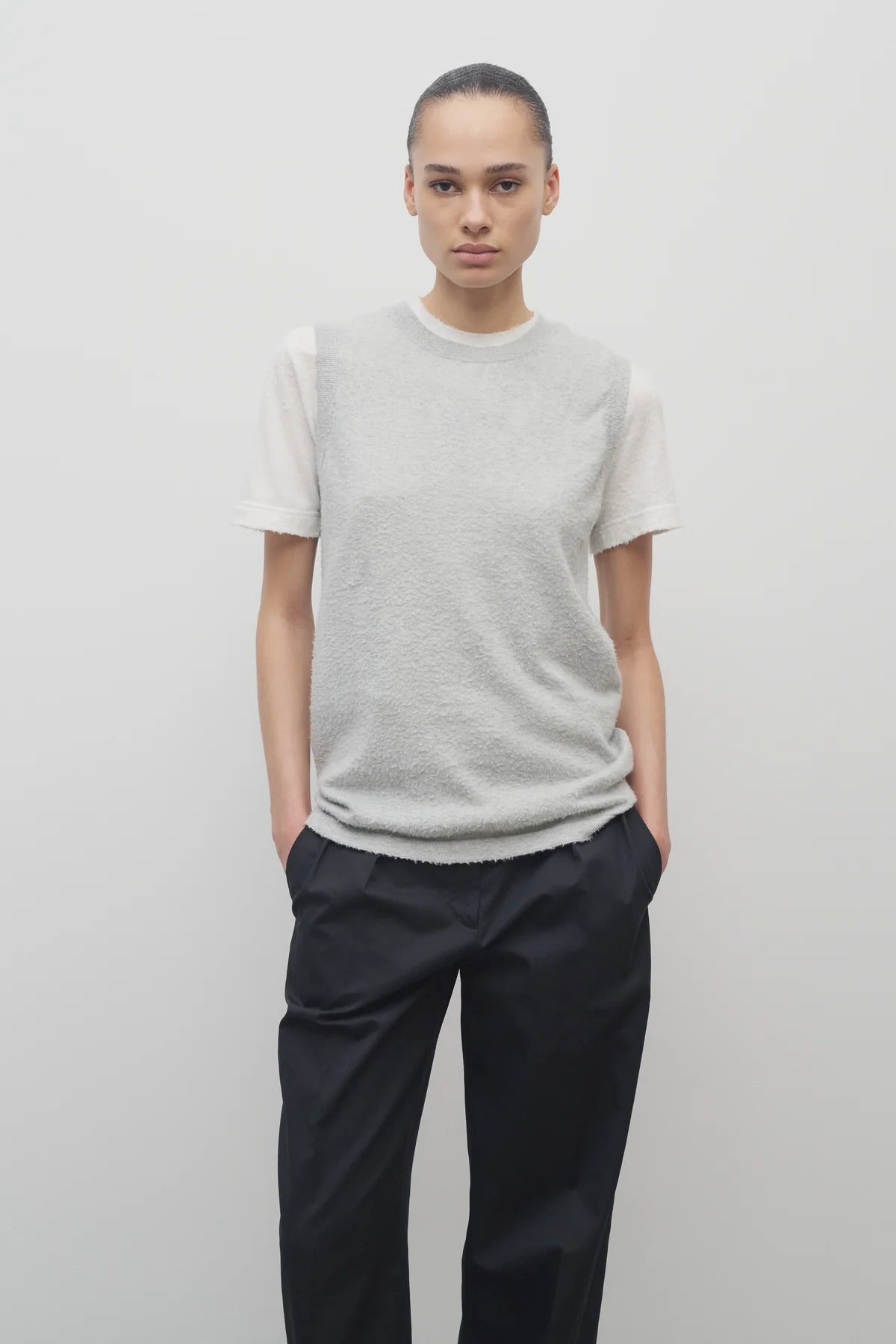 A model with dark brown hair is shown in the sleeveless knit top and navy trousers with frontside viewpoint on white backdrop
