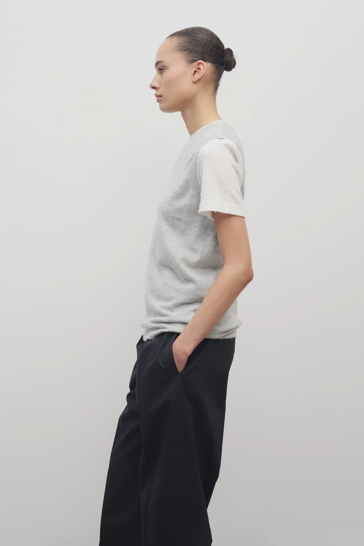 A model with dark brown hair wears the sleeveless knit top in grey melange worn with navy trousers shown at a side angle against a white backdrop